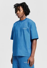 Sininen puuvillapaita, jossa on lyhyet hihat, rento istuvuus ja pyöreä pääntie. Paidan rinnassa on brodeerattu teksti, ja se on yhdistetty samansävyisiin shortsit.