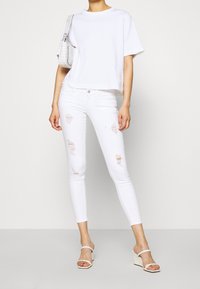 Camiseta blanca de manga corta combinada con jeans ajustados rotos blancos. La modelo lleva sandalias de cuña blancas y sostiene un bolso texturizado blanco.