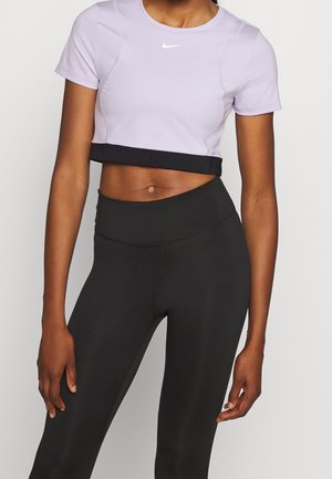 Femme portant un t-shirt de sport court Nike lavande et des leggings noirs taille haute, debout sur un fond blanc uni.