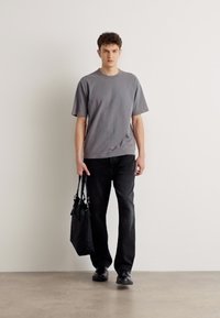 T-shirt gris à col rond, coupe ample, associé à un jean noir à jambes droites. Le mannequin tient un sac à bandoulière noir, avec des chaussures noires �élégantes.