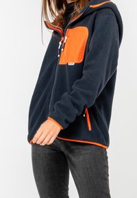 Merenneito fleece-takki, jossa on oranssi rintatasku ja yksityiskohtia, edessä vetoketju, huppu ja joustavat hihansuut. Teksturoitu kangas, jossa on rento istuvuus.