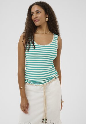 Femeie cu păr creț, purtând un top fără mâneci în dungi turcoaz și alb și o fustă albă cu o curea împletită și ciucuri.