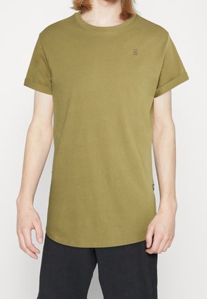 Person iført olivengrøn kortærmet T-shirt med lille logo på brystet og sorte ribbukser mod ensfarvet baggrund.