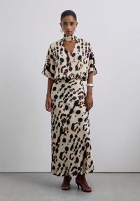 Mujer vistiendo un vestido maxi de color beige y negro con manchas, con un escote de gargantilla, pendientes de aro plateados, pulsera y tacones negros con tiras, de pie.
