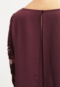 Vue arrière d'un chemisier bordeaux avec une ouverture en forme de trou de serrure et une fermeture à bouton au niveau de l'encolure, avec des manches en dentelle.