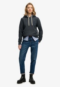 Sudadera con capucha gris oscuro con un logo, cordones y bolsillo, combinada con una camisa a rayas azules y blancas. Combinada con vaqueros azules y botas negras.