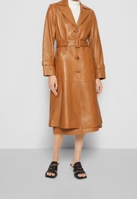 Trench-coat en cuir marron avec une taille ceinturée, de grands revers, des boutons et des manches longues, associé à des sandales noires à brides.