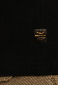 Crna tkanina majica s rektangularnom žutom i crnom etiketom na kojoj se nalaze natpisi "PME LEGEND" i "AMERICAN CLASSIC", iznad bež kordurovskog tekstura.
