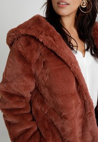 Dirbtinio kailio paltas rudų atspalvių, su švelniu, tekstūruotu paviršiumi. Turi gobtuvą ir atvirą priekinį dizainą, dėvimas ant baltos bliuskos.