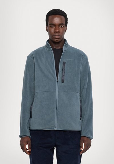 Veste en polaire teal avec un col haut, une fermeture éclair à l'avant et une poche poitrine avec un accent noir contrastant. Texture lisse.