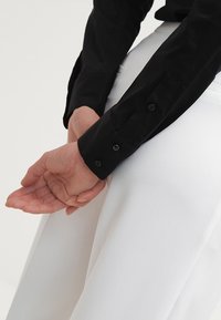 Czarna koszula z długim rękawem i mankietami na guziki, noszona z białą spódnicą. Koszula ma gładką teksturę i dopasowany krój.