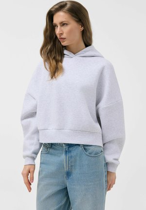 Kobieta w jasnoszarej oversize bluzie z kapturem i jasnoniebieskich dżinsach, patrząca w bok na jednolitym tle.