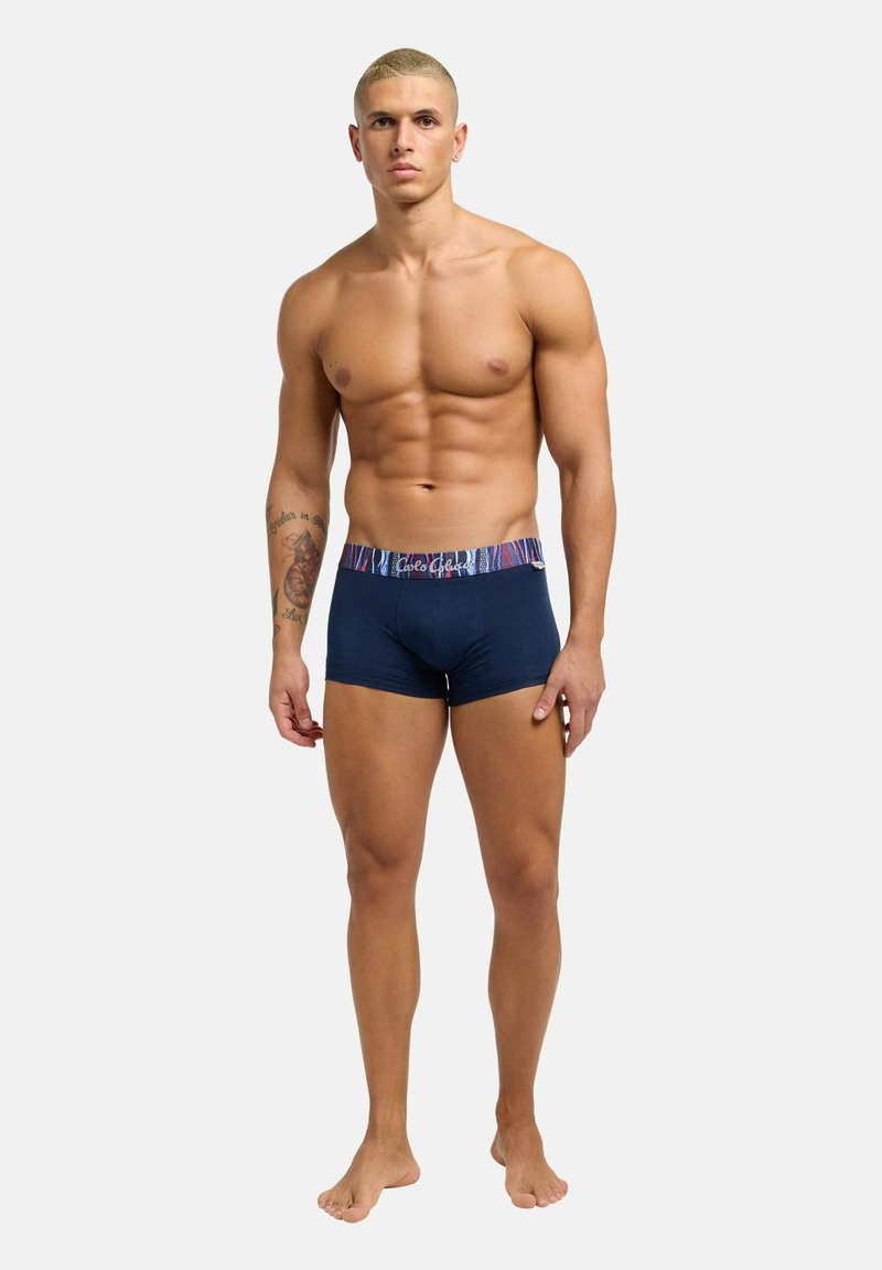 Muscular man with short blond hair stands barefoot wearing navy blue boxer briefs with patterned waistband, neutral expression, white background.