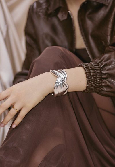 Femme portant un large bracelet manchette torsadé en argent, une bague en or, une veste en cuir marron et une jupe marron transparente, le bras reposant sur le genou.