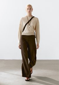 Maglione a maglia beige e pantaloni a gamba larga marrone scuro. L'outfit è abbinato a sandali neri. Una collana con cinturino nero e pendente in argento aggiunge un tocco di dettaglio.