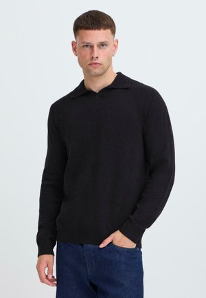 Zwarte gebreide trui met een colkraag, lange mouwen en geribde manchetten. Draag het met blauwe denim broek, weergegeven tegen een lichte achtergrond.