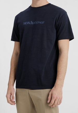 Mężczyzna w granatowym t-shircie z napisem „w.w.hatever” i beżowych spodniach, stoi z rękami swobodnie opuszczonymi wzdłuż ciała na jednolitym tle.