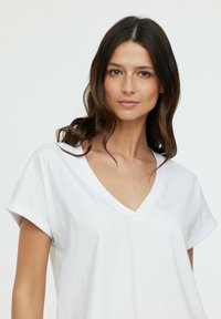 T-shirt en coton blanc avec un col en V, manches courtes retroussées et coupe décontractée. Texture lisse, design simple, sans motifs ni embellissements.