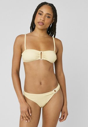 Femme avec des cheveux tressés portant un bikini jaune pâle avec un détail en anneau sur le bas, debout devant un fond gris neutre.