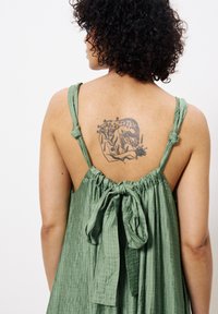 Grünes, gekräuseltes Kleid aus Stoff mit Bindebandträgern und gerafftem Bund, das ein Tattoo einer Figur und Pflanzen auf dem oberen Rücken der Trägerin zeigt.
