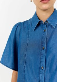 Niebieska koszula dżinsowa z krótkimi puffowanymi rękawami, zapięciem na guziki z przodu i wyraźnym kołnierzykiem. Posiada pionowe szwy i kontrastowe brązowe guziki.