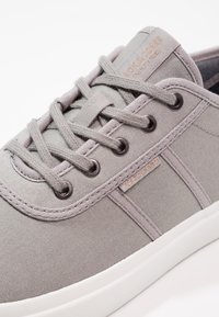 Baskets en toile grise avec bout arrondi, lacets plats et œillets métalliques. Comprend une étiquette logo et une semelle texturée contrastant en blanc.