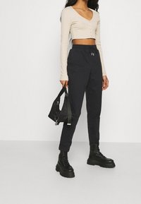 Beżowy krótki top z długim rękawem zapinany na guziki, zestawiony z czarnymi zwężającymi się dresami i masywnymi czarnymi butami na sznurówki, trzymający małą czarną torbę.