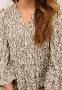 Femme portant une robe marbrée crème et marron avec un décolleté en V et de longues manches bouffantes, les cheveux tombant sur son épaule.