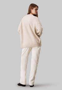 Beige oversized trui van zacht materiaal met een geribbelde zoom, gecombineerd met witte corduroy broek en zwarte enkelboots.