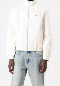 White lightweight zip-up jacket with high collar, elastic cuffs, and hem; small logo detail on the chest; worn over light blue jeans.