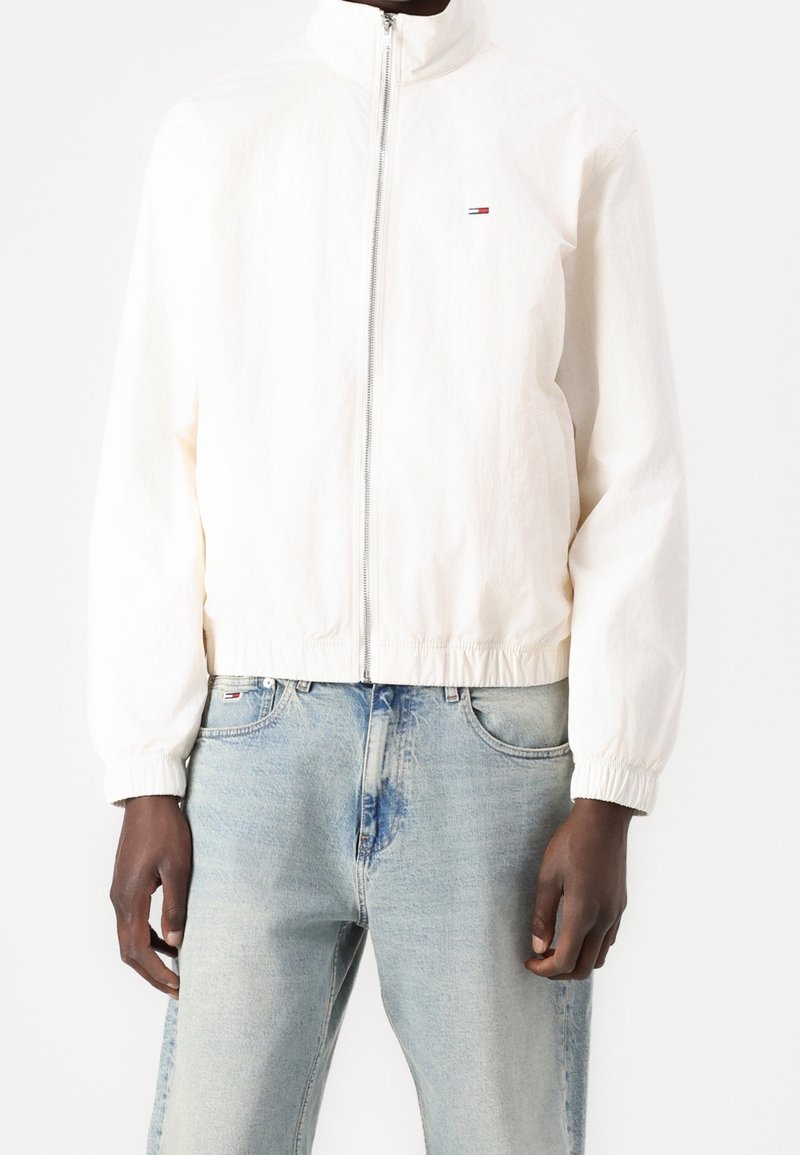 White lightweight zip-up jacket with high collar, elastic cuffs, and hem; small logo detail on the chest; worn over light blue jeans.