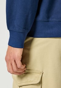 Niebieska bluza z ribbanymi mankietami, wyeksponowana z ręką spoczywającą na beżowych spodniach cargo z kieszenią na klapkę.