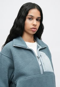 Νεαρός με μακριά κυματιστά μαύρα μαλλιά, φορώντας μπλε fleece μπουφάν με φερμουάρ στη θηλειά, που στέκεται μπροστά σε μια απλή λευκή επιφάνεια.