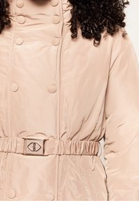 Beige vadderad jacka med tryckknappar, elastisk midja och en metallspänne-detalj. Slät textur, hög krage och sidofickor.