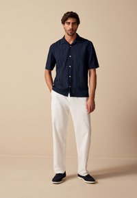 Kortærmet navyblå skjorte med lodrette striber, knaplukning og spredt krave, kombineret med hvide linnedbukser og navy slip-on sko.