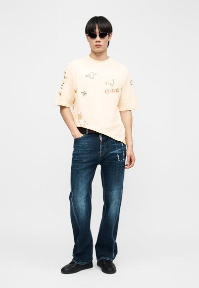Άνδρας με μαύρα γυαλιά ηλίου, κρεμ μπλουζάκι με στάμπα, σκούρο μπλε τζιν και μαύρα παπούτσια, στέκεται με το ένα χέρι στην τσέπη.