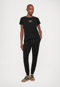 T-shirt preta com logótipo dourado, combinada com calças de fato pretas e ténis verdes. Apresenta um corte descontraído e um design simples.