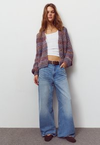 Cardigan tricotat din fire multicolore, cu nasturi în față, asortat cu blugi largi, de culoare albastru deschis, și o curea maro.