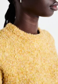 Texturerad gul tröja med rund halsringning. Tyget verkar mjukt och har en fluffig yta med inslag av rosa här och där.