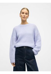 Maglione oversize color lilla chiaro con polsini e scollo a coste, abbinato a pantaloni in denim scuro con cuciture visibili e tasche laterali.