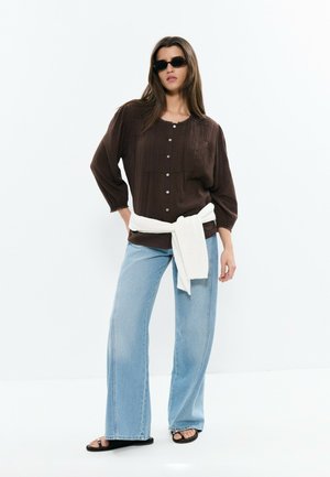 Mujer con blusa marrón oscuro abotonada, jeans de pierna ancha azul claro, sandalias negras, suéter blanco atado a la cintura y gafas de sol oscuras.