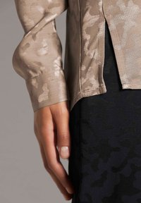 Beige Satin-Oberteil mit langen Ärmeln und subtilen Camouflage-Muster, mit einem seitlichen Schlitz. Schwarzer strukturierter Unterteil teilweise sichtbar darunter.