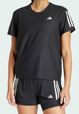 Kobieta ubrana w czarną odzież sportową Adidas z białymi paskami na rękawach i szortach, stojąca na jasnym tle.