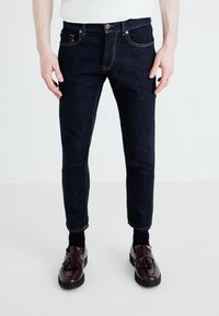 Tmavě modré džínové kalhoty s oranžovým prošíváním, slim fit designem a klasickými kapsami, doplněné o tmavé boty. Textura vypadá hladce.