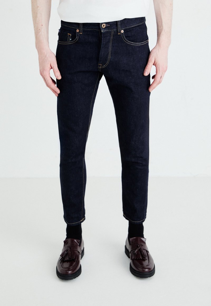 Tmavě modré džínové kalhoty s oranžovým prošíváním, slim fit designem a klasickými kapsami, doplněné o tmavé boty. Textura vypadá hladce.