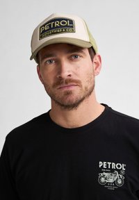 Gorra con un panel frontal crema y parte trasera de malla verde oliva, que presenta el logo "PETROL INDUSTRIES & CO." Camiseta negra con un gráfico de motocicleta.