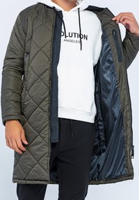 Olivengrøn quiltet lang frakke med lynlåslukning, der har en kontrasterende indvendig foring og en sidelomme. Glat ydre tekstur.