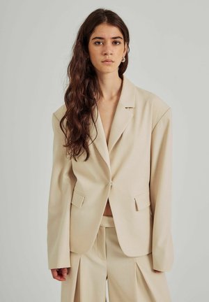 Beige sakko na míru s jedním knoflíkem, klopovými límci a bočními kapsami. Tkanina má hladkou strukturu a mírně tvarovaný střih.