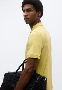 Κίτρινο πόλο με κοντά μανίκια, συνδυασμένο με μια μαύρη δερμάτινη τσάντα με υφή και διπλές λαβές. Απλός, σύγχρονος σχεδιασμός.