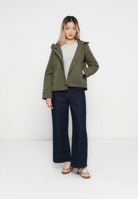 Veste à capuche zippée vert olive avec des boutons-pression ; portée sur un haut gris clair et associée à un jean bleu foncé à jambes larges et des baskets beiges.